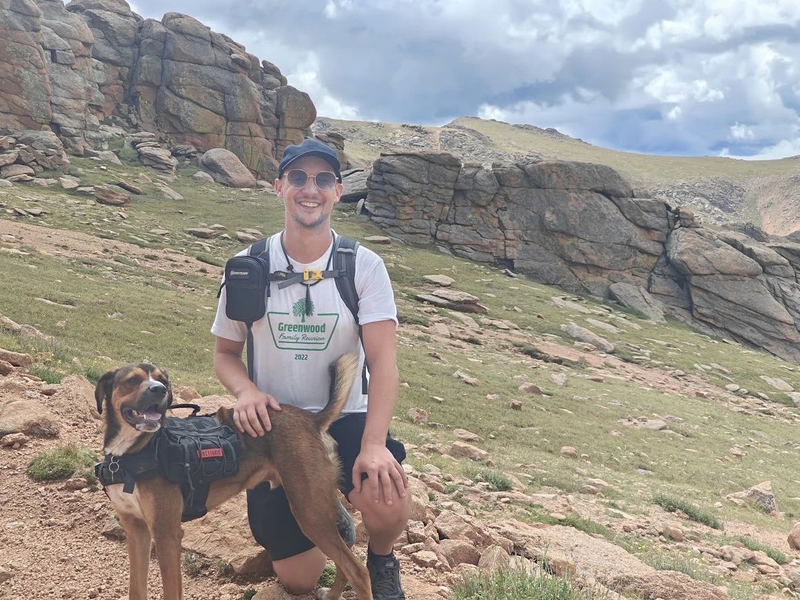 Eric kneeling on a rocky mountain trail with his dog, Bruno, both smiling